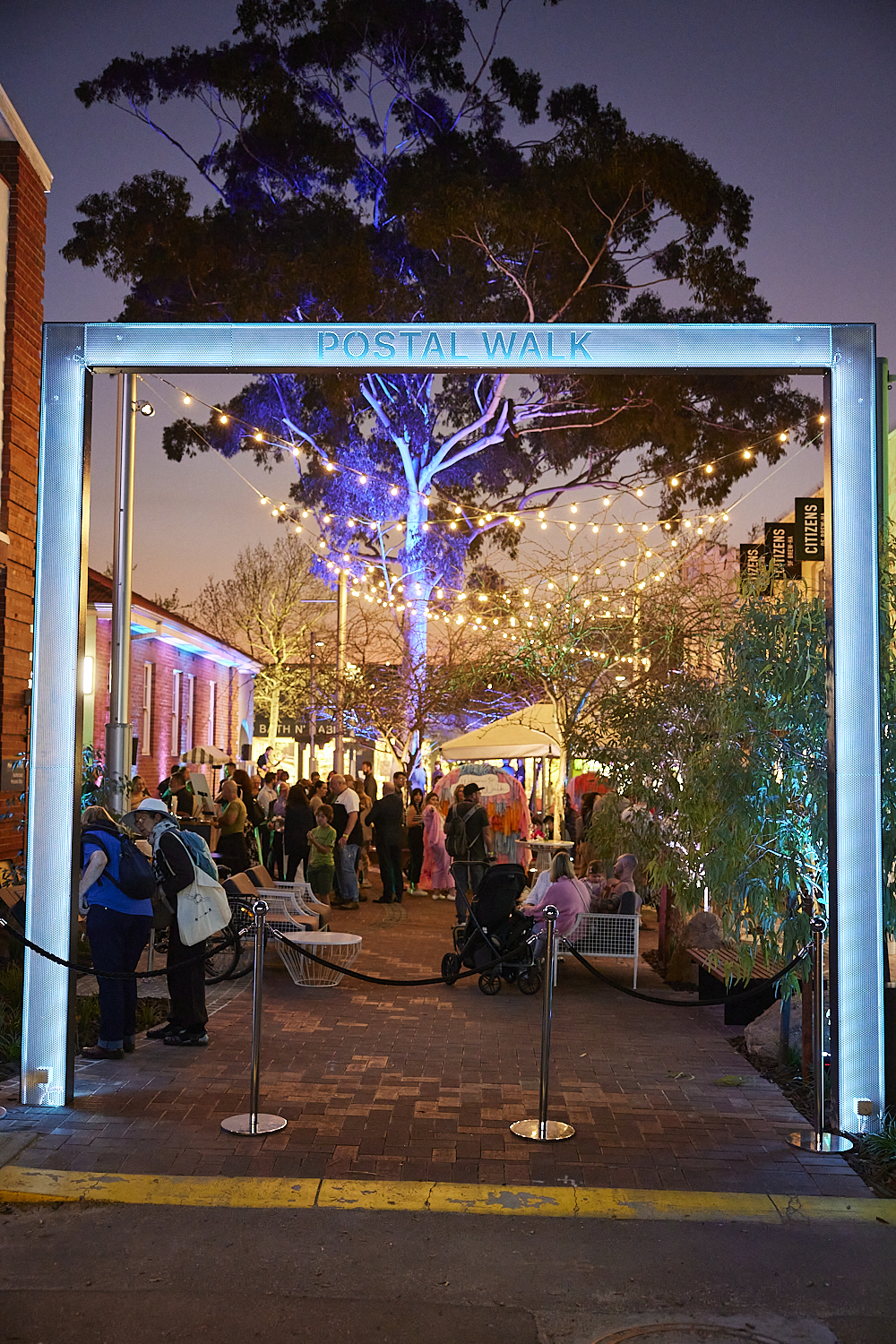 People celebrating in a brightly lit, newly upgraded public space. Fairy lights hang in the tree canopy overhead.